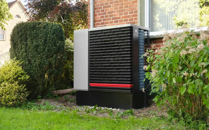 Pompe à chaleur air-eau installée dans le jardin d'une maison en briques, entre des arbustes